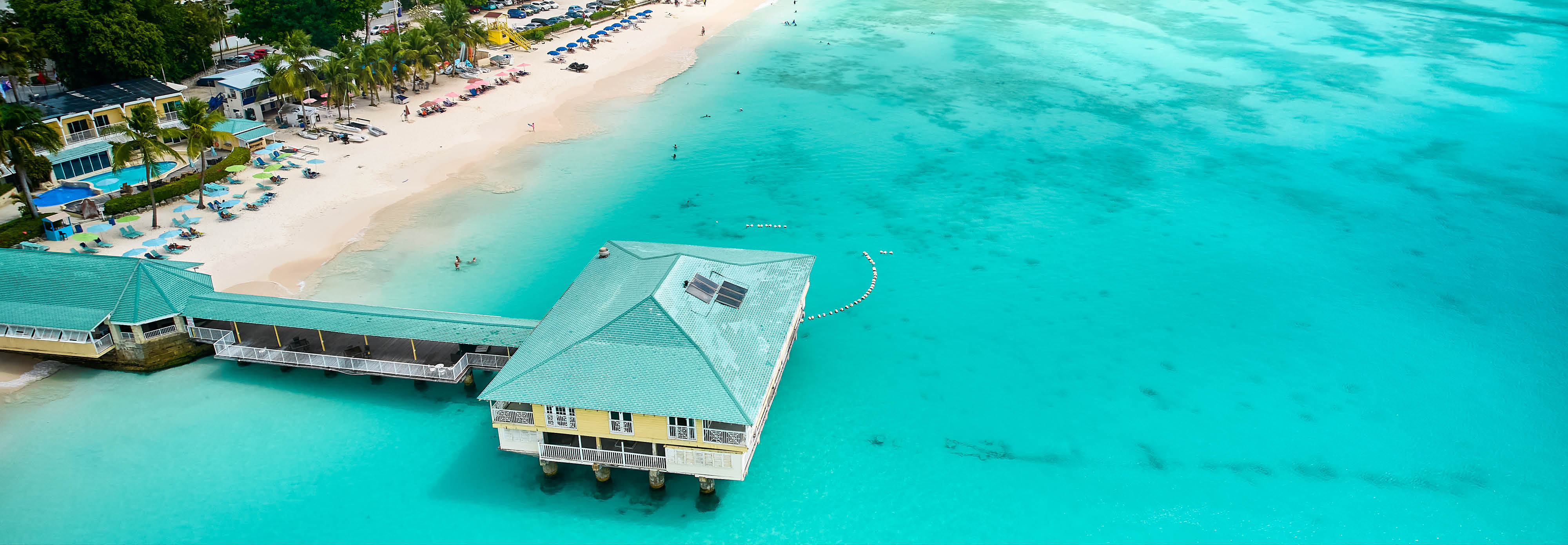 Aerial view of a sandy beach with a long pier extending into the water, surrounded by beachgoers in Barbados