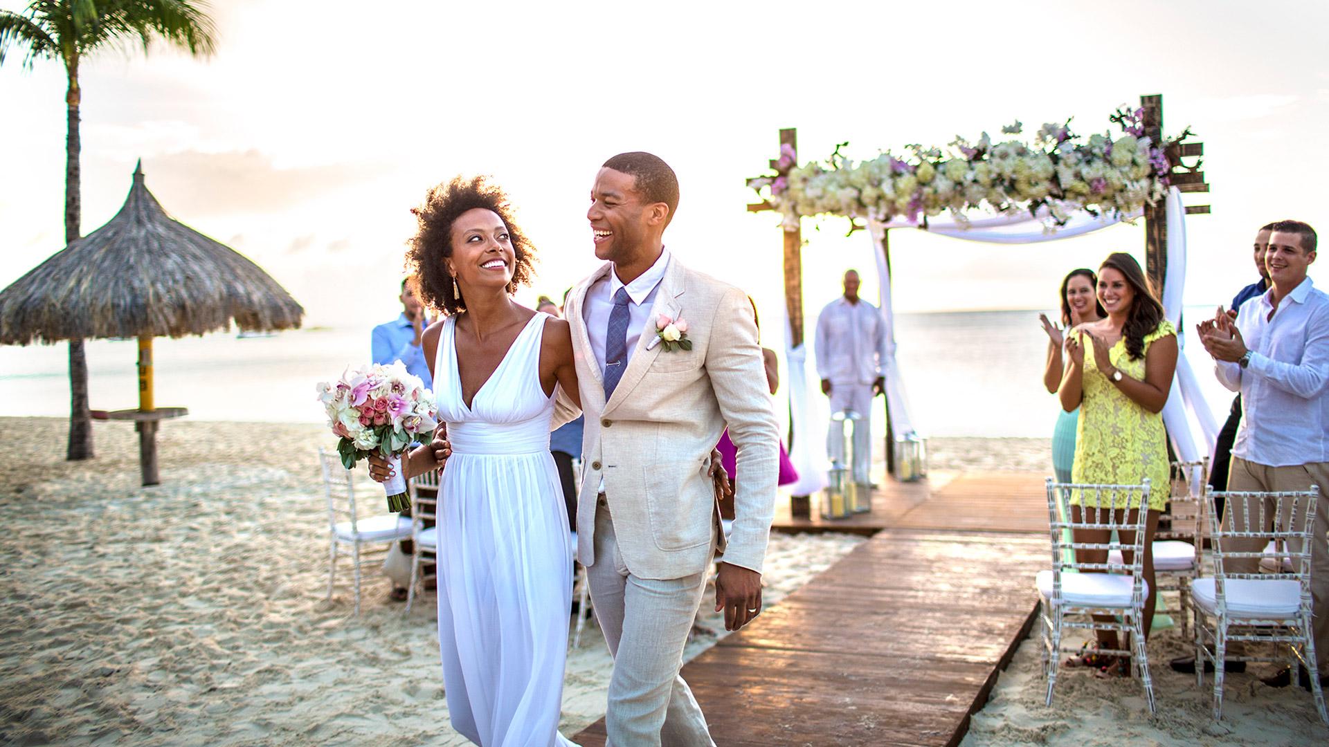 A joyful couple walks arm in arm down a sandy beach aisle after their wedding ceremony, surrounded by smiling and clapping guests under a floral arch at sunset.