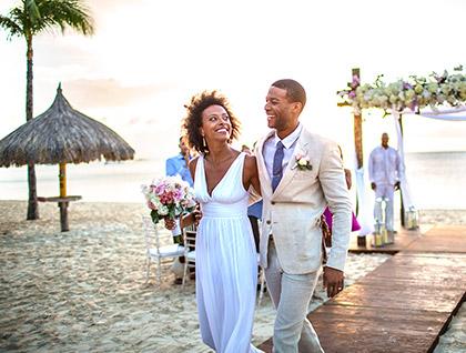 A joyful couple walks arm in arm down a sandy beach aisle after their wedding ceremony, surrounded by smiling and clapping guests under a floral arch at sunset.