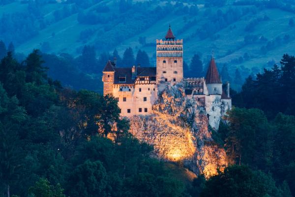 Lit-up castle on a rocky hill surrounded by trees at dusk.