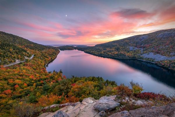Pink sunset sky over a lake surrounded by autumn trees and mountains.