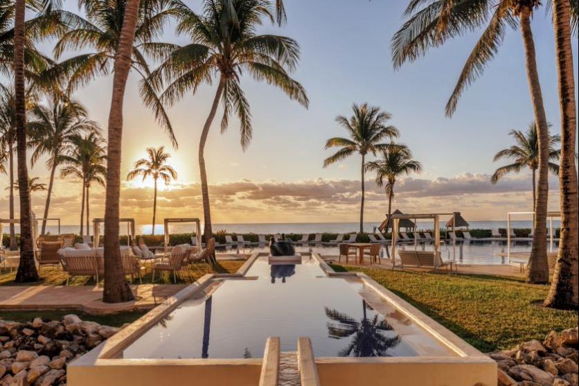 A luxury Royalton resort pool area at sunset, framed by palm trees, with a long reflecting pool leading to the ocean and lounge chairs.