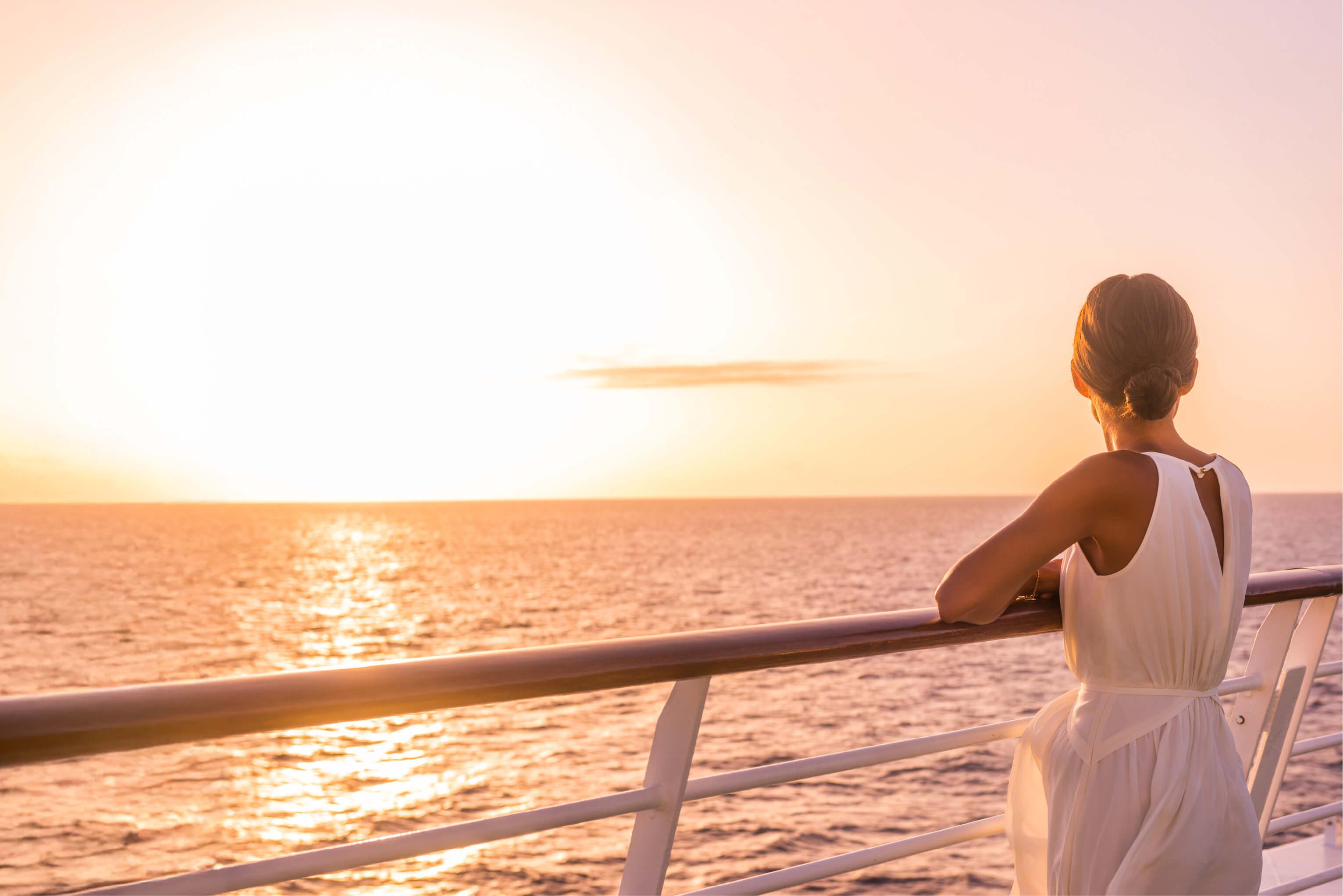 A woman in a white dress stands on the balcony of cruise ship staring out at the sunset over the ocean