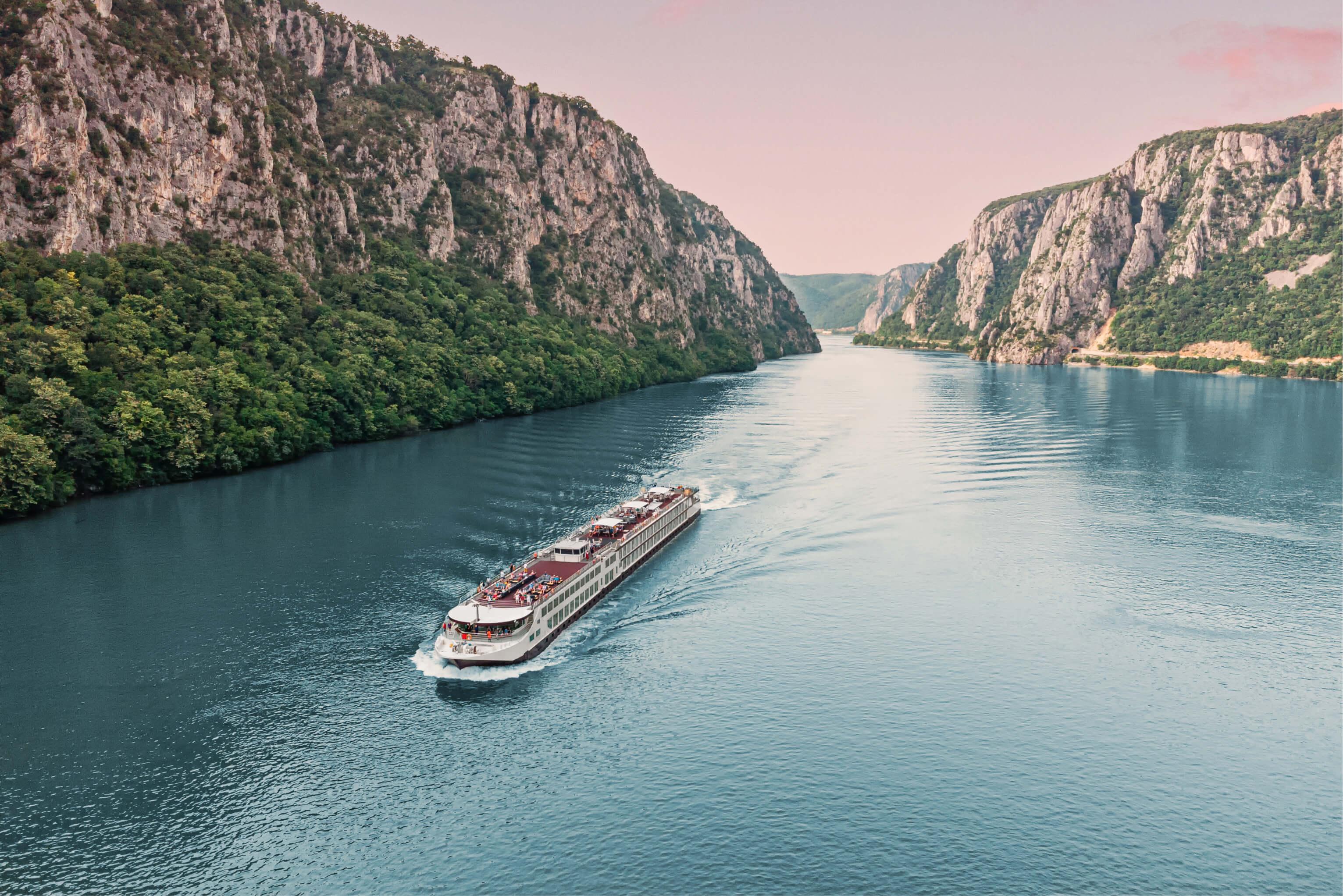 A cruise ship navigates a wide river toward a pass between two mountains