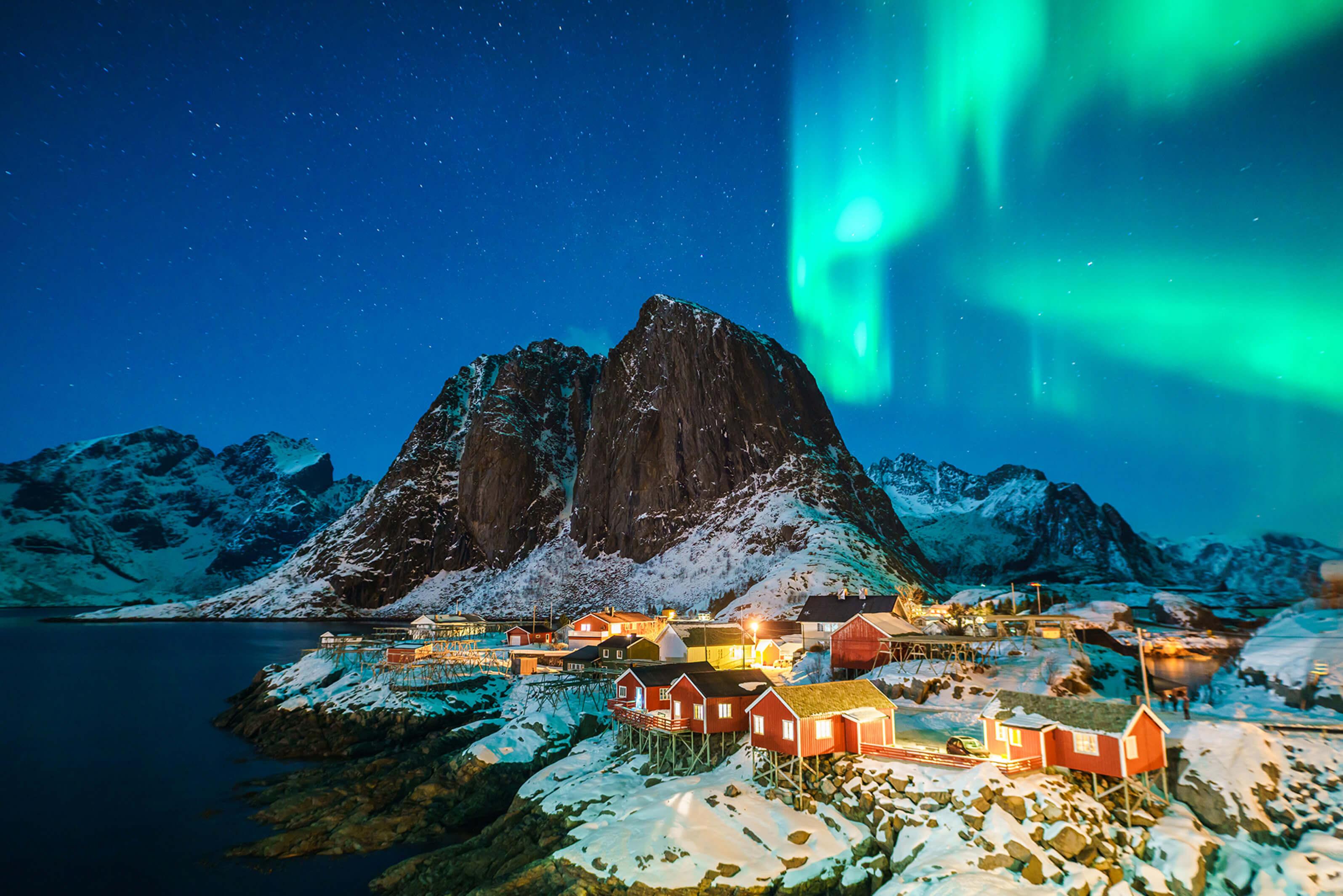 The aurora borealis illuminates the night sky above a mountain and a nearby village in Iceland, creating a stunning natural display