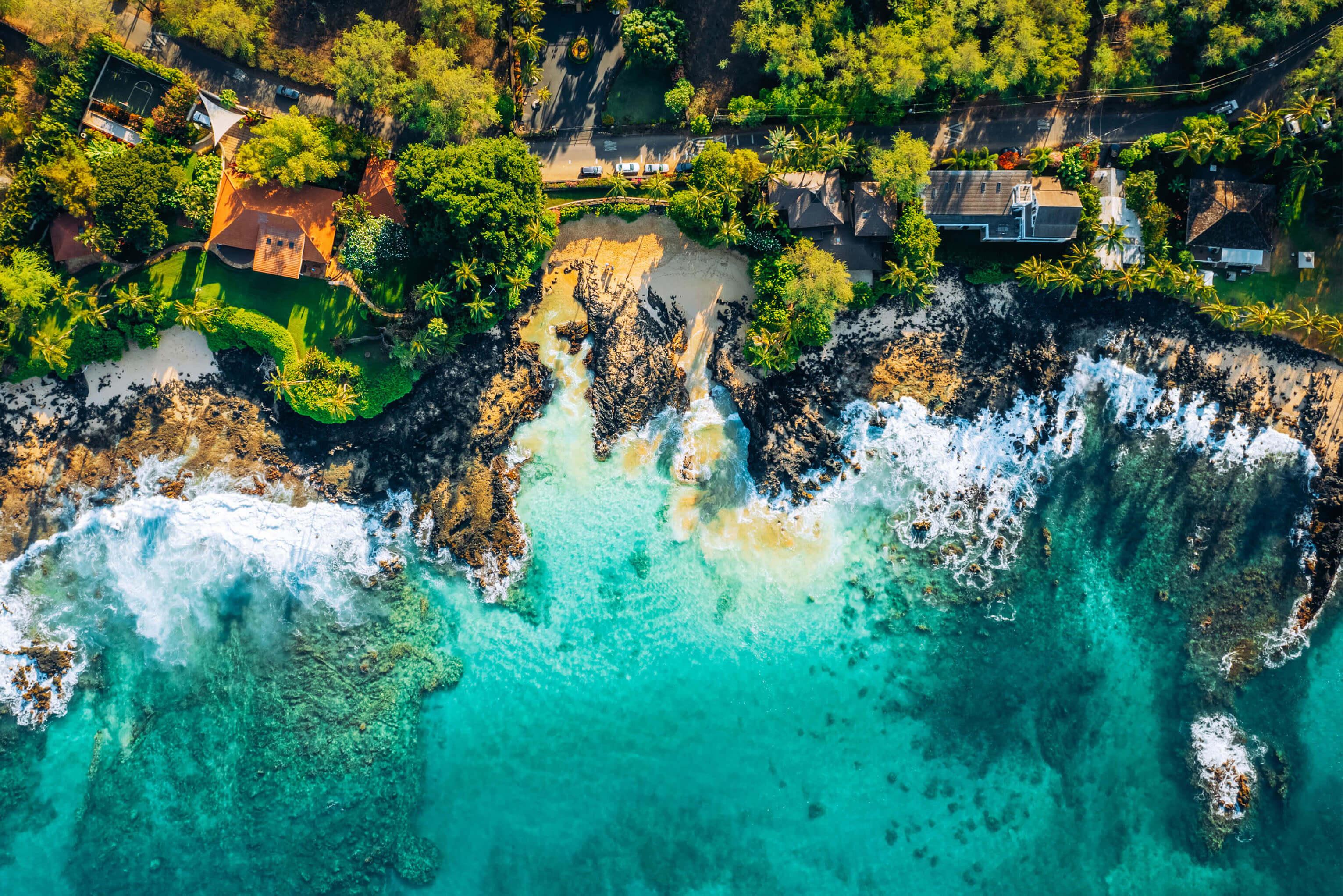 Overhead view of a Hawaiian beach in Maui with golden sand and residential homes nestled among palm trees and vibrant foliage