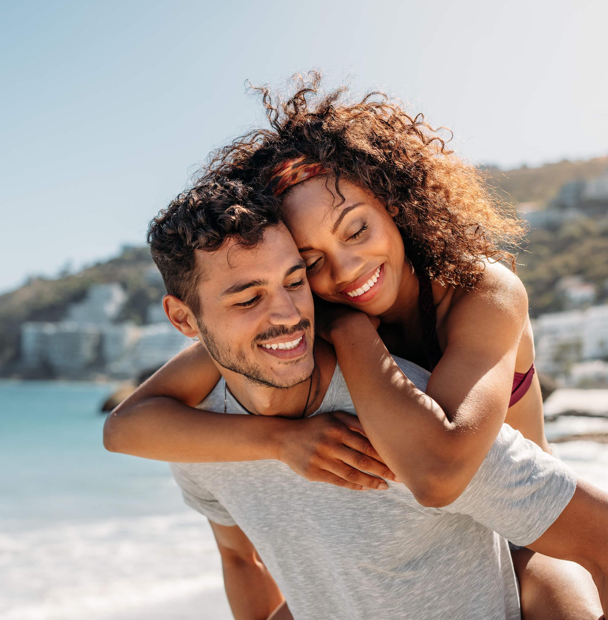 Happy couple enjoying a romantic beach escape, with a man carrying his partner along the shoreline during a sunny holiday.