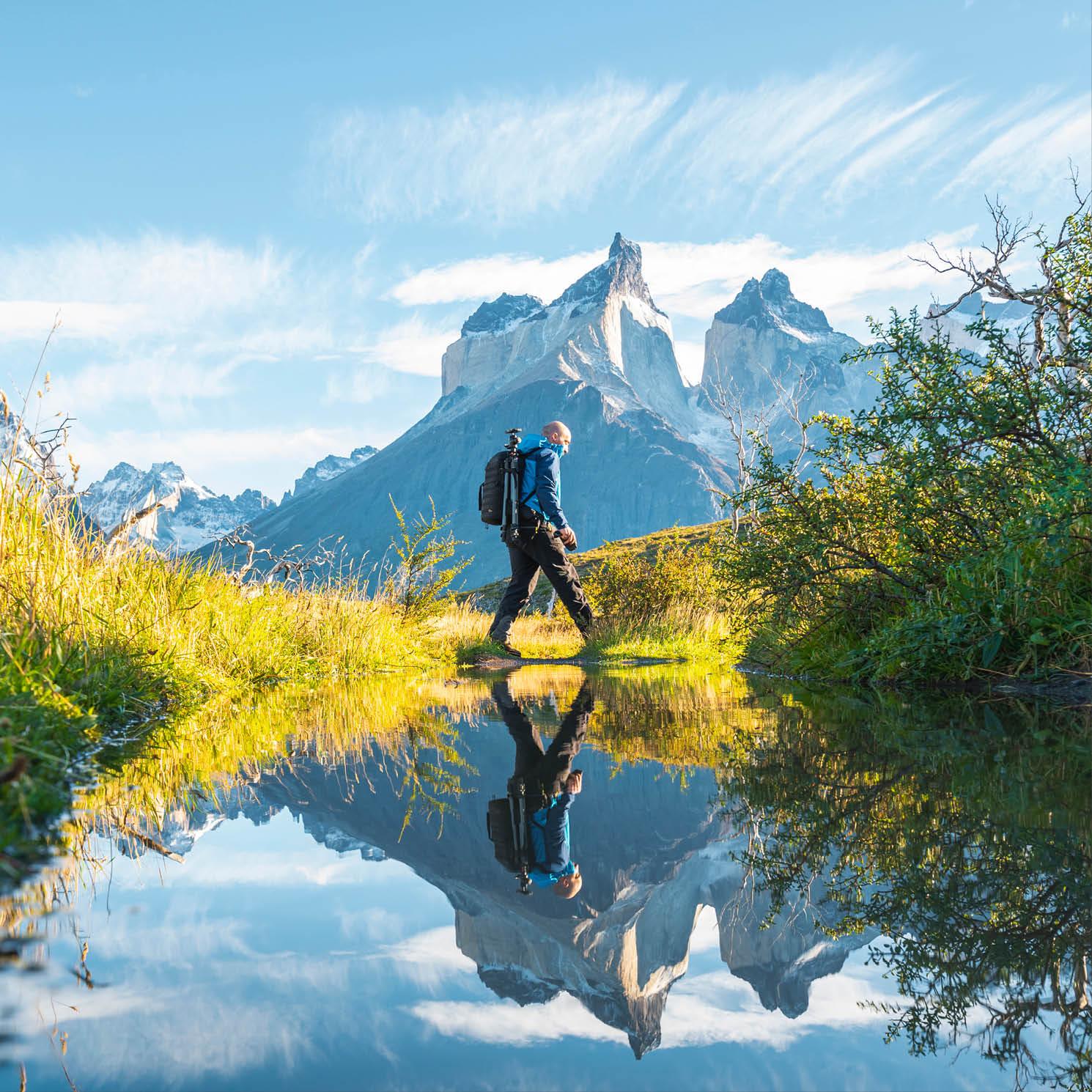 A hiker crosses a body of water, with his reflection visible and mountains in the background